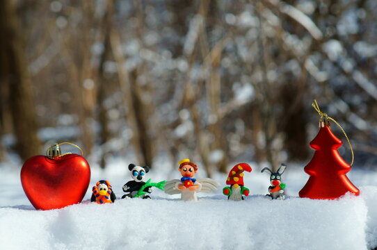 Figurines of fairy-tale characters in the winter forest. An angel with a gift, a dwarf, a rabbit with a carrot, a panda, a penguin in a scarf. Next to the Christmas tree and a red heart.