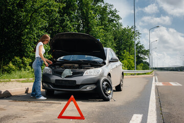 A young girl stands near a broken-down car in the middle of the highway and looks under the hood....