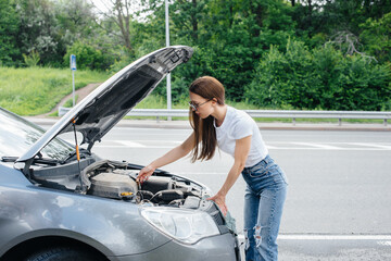 A young girl stands near a broken-down car in the middle of the highway and looks under the hood....