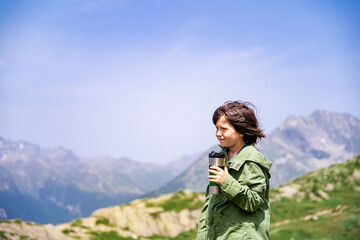 Young boy of ten years old staying in a mountains, holding travel tumbler and drinking tea. Child hiking in the highlands.  Kid looking aside and admiring wild nature.