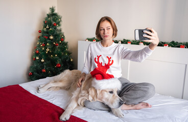Obraz premium Portre of a young woman with a dog in a Christmas atmosphere. The girl looks at the phone in the hat of Santa Claus with a golden retriever on the bed near the Christmas tree.