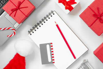 Christmas online shopping flat lay. Notebook with pencil. Shopping list. Credit card, Laptop, Candy and Christmas presents on white table. Page for your write. Copy Space. Winter holidays sales
