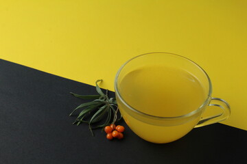 sea buckthorn tea drink on a black yellow bright background. Sea buckthorn drink tea juice in a glass cup next to a spout of sea buckthorn leaves orange berries on a black and yellow background. 