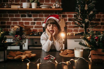 a dreaming little boy is sitting at the kitchen table in a white sweater and a red Christmas hat