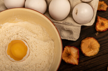 A broken white chicken egg in a bowl with wheat flour, dried apple chips and several whole eggs in a tray made of white cardboard, close-up selective focus.