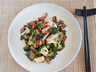 top view of stir fry on a plate with vegetables, prowns and meat. blck chopsticks on a bamboo tablecloth