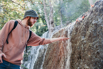 Joven barbudo con un sombrero de pescador tocando el agua que corre por la pared de una cascada