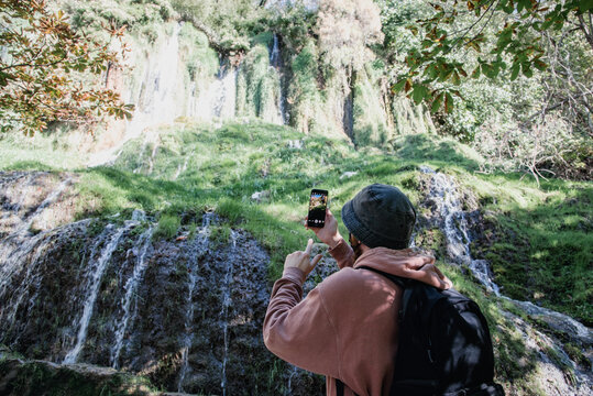 Hombre joven con un bucket hat tomando una foto con su tel&eacute;fono m&oacute;vil frente a una cascada