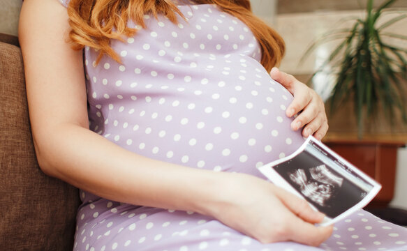 pregnant young woman holding ultrasound picture sitting in kitchen, happy pregnancy concept