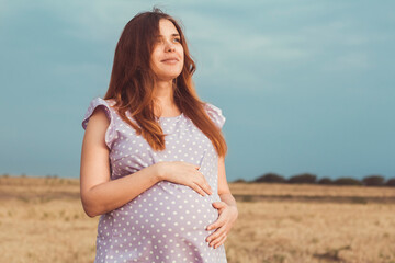 beautiful red-haired pregnant woman in summer dress stands against background of rainy gloomy sky with clouds, happy girl in anticipation of child in field