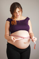 beautiful red-haired pregnant woman use tape measure at big belly on gray studio background, female body during pregnancy