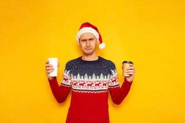 sad, dissatisfied, disappointed man in Christmas sweater with reindeer red Santa Claus hat holds paper cups with hot drinks tea, coffee. isolated yellow background with space for text concept new year