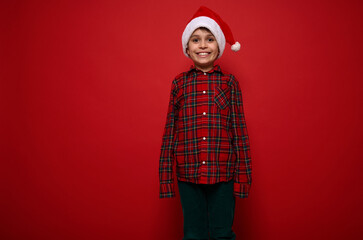 Fototapeta premium Isolated portrait on colored background with copy space for Christmas and New Year ad of cheerful funny cute preadolescent boy in red plaid shirt and green velvet trousers smiling looking at camera