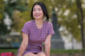 Naklejka premium urban lifestyle outdoors portrait of young happy and beautiful Asian Korean woman in sweet Autumn dress enjoying Madrid city tour relaxed and smiling sitting on park bench