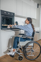 A girl on a wheelchair opening oven in the kitchen while cooking soemthing