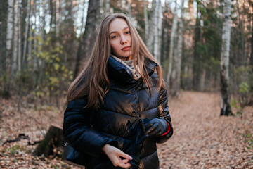 Smiling young woman in coat and scarf standing in autumn forest. Woman looking at the sunset on trees background and breathing fresh air. Person in nature. Having fun outdoors.