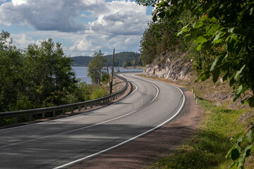 Curve of an empty asphalt countryside highway. The road has 2 lanes, fenced and with white markings. Large stones, rocks and trees is on the side of the road, on the other side there is a Ladoga lake.
