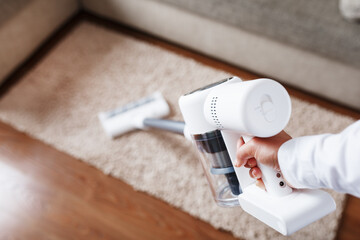 Powerful cordless vacuum cleaner with white cyclonic dust collection technology in hand, cleans the carpet in the house near the sofa. - obrazy, fototapety, plakaty