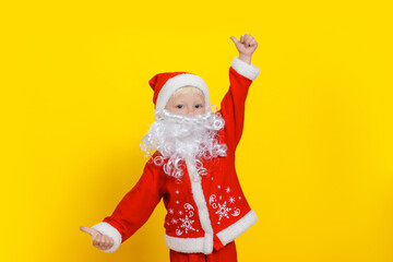 Three-year-old child in Santa Claus clothes and with a white beard shows a thumbs up gesture, stands on a yellow studio background.