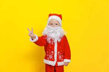 Fototapeta na wymiar Little boy in red christmas costume with white beard shows victory gesture with hands, stands on yellow isolated background.