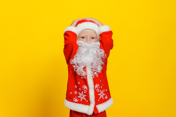 Caucasian child in New Year Santa Claus costume and white artificial beard holding his head, yellow isolated background.