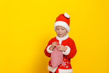 A small child dressed in a red Santa Claus costume is holding a small sack with New Year's gifts. Yellow studio background.