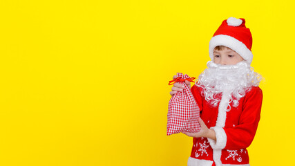 A child in Santa Claus clothes and an artificial beard holds a small bag with New Year's gifts in his hands, copy space, yellow studio background.