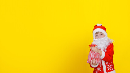 Little caucasian boy in Santa Claus clothes and an artificial white beard holds a sack with New Year's gifts in his hands, looks at the camera, copy space, yellow studio background.