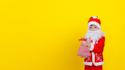 A little boy in Santa Claus clothes and an artificial beard holds a small bag with New Year's gifts in his hands, looks at the camera, copy space, yellow studio background.