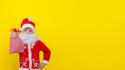 Fototapeta na wymiar A six-year-old Caucasian child in Santa Claus clothes and with an artificial white beard shows a bag of gifts, looks at the camera, copy space, yellow studio background.