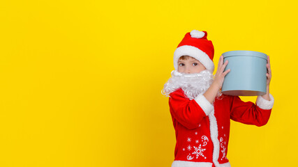 Cute Caucasian child in Santa Claus clothes and with an artificial white beard holding a round gift box high in his hands, yellow studio background.