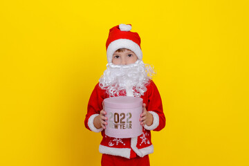 A cute Caucasian boy in Santa Claus clothes and with an artificial white beard holds a pink round gift box with the words New Year 2022 in his hands, yellow studio background.