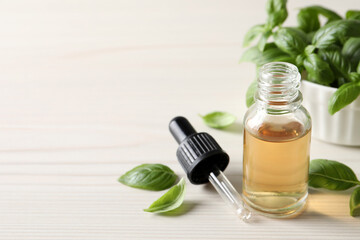 Glass bottle of basil essential oil and leaves on white wooden table. Space for text