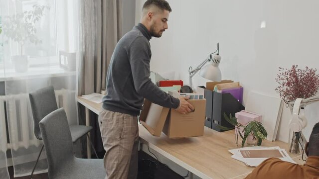 Tracking medium shot of young male office worker packing his personal belongings into cardboard box while leaving office. His colleagues continue working on computers