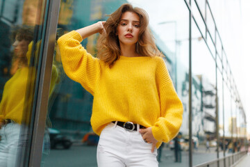 Front image of a young woman with brown wavy hair, wear in yellow sweater and white jeans behind modern building.