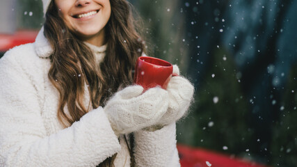 Happy beautiful young woman wearing knitted sweater and woolen hat drinking hot chocolate near red...