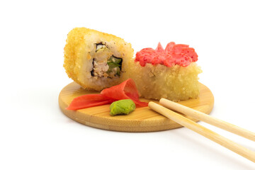 Still life with hot fried sushi rolls on round wooden plate with chopsticks, pickled pink ginger and wasabi isolated on white background. Cheap tempura roll with chicken, cucumber and pink mayonnaise