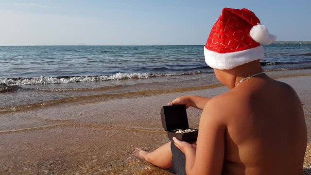 Santa Claus in swimming trunks and a New Year's hat is sitting on the beach by the sea and looking at a casket with sea gifts. Christmas on the beach of ocean waves