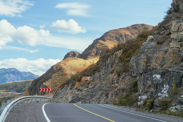 Colorful autumn landscape with mountain highway along rocks in sunshine. Bright alpine scenery with mountain road in autumn colors. Highway in mountains in fall time. Road along beautiful rocky wall.