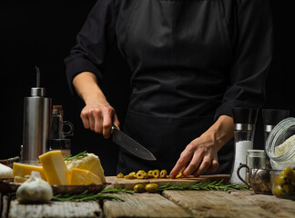 The chef is cutting olives on a cutting board. Ingredients for making salad, pizza. ravioli, pasta. Wooden texture, black background. A restaurant. hotel, recipe book, cookbook.