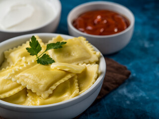 On a dark blue background, a portion of Italian ravioli with a mint leaf. Sour cream and tomato sauces. Brown napkin, blue background. Close-up. Cookbook, culinary blog, menu design.