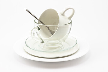 Balanced tower from transparent glass and white ceramic tableware isolated on white background. Pile of tea cups and plates with metal silver spoon