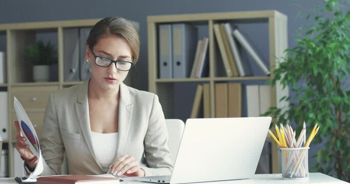 Young beautiful female office manager sitting at workplace and focused working with paper documents checking infographics on laptop.