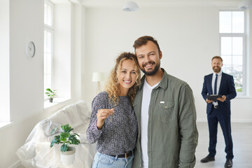 Happy smiling young married couple looking at camera and showing key to new home standing in...
