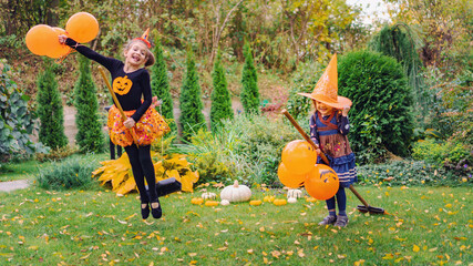 Children in witch hats laugh and play with brooms. Little witches learn to fly broomsticks with orange balloons at an outdoor Halloween party in the fall season. Fun ideas for kids at Halloween party.