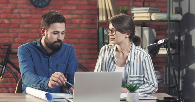 Two young caucasisn colleagues, man and woman, sit together at desk in modern office and brainstorm over joint project.