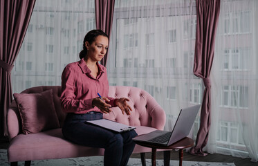 a Caucasian female psychologist in a pink shirt sits on the couch and conducts an online consultation on a laptop
