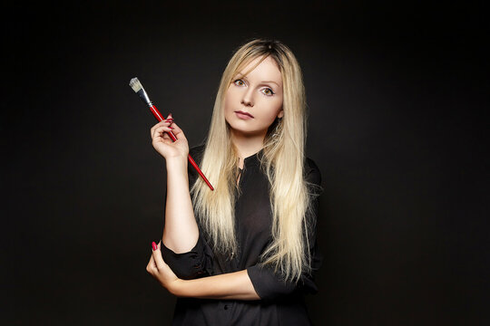young blonde artist poses with a brush in her hand. photo shoot on a black background in the studio