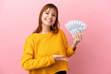 Redhead girl taking a lot of money isolated on pink background extending hands to the side for inviting to come