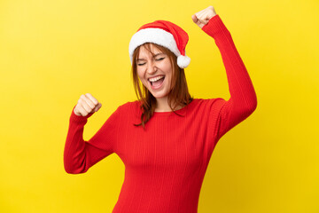 Redhead girl with christmas hat isolated on yellow background celebrating a victory